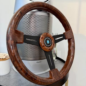 Puede incluir: Un volante de madera con acabado marrón oscuro y radios negros. El centro presenta un emblema circular con el texto "NARDI". El volante está diseñado para un vehículo y tiene una apariencia clásica y elegante.