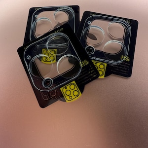 以下が含まれることがあります： スマートフォン用の透明なプラスチック製カメラレンズ保護フィルム3枚。保護フィルムはピンクの表面に置かれています。保護フィルムは黒い縁取りと透明な中心部があります。保護フィルムは、カメラレンズを傷や損傷から保護するために設計されています。