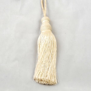 May include: A cream-coloured decorative tassel featuring a tightly wound cord loop at the top, a structured knot section, and a long, flowing fringe made of fine, silky threads that hang straight down in a uniform, vertical arrangement.