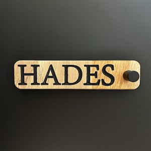 Puede incluir: Un letrero de madera con la palabra "HADES" tallada en letras negras. El letrero tiene un pomo negro en el lado derecho.
