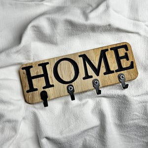 Puede incluir: Porta llaves de madera con la palabra "HOME" en letras negras y cinco ganchos de metal negro.