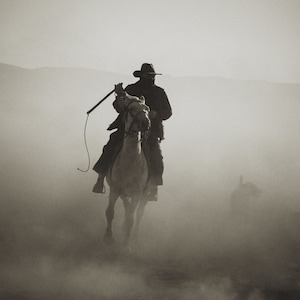 Puede incluir: Un vaquero montando un caballo blanco a trav&eacute;s de un paisaje polvoriento. El vaquero lleva un sombrero negro y una gabardina larga. Sostiene un l&aacute;tigo en su mano.