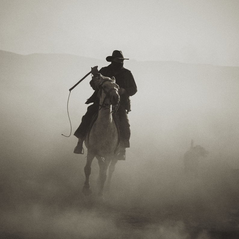 Puede incluir: Un vaquero montando un caballo blanco a trav&eacute;s de un paisaje polvoriento. El vaquero lleva un sombrero negro y una gabardina larga. Sostiene un l&aacute;tigo en su mano.