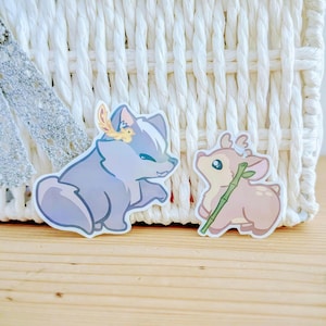 Peut inclure: Deux autocollants représentant des animaux de dessin animé. L'un est une créature ressemblant à un renard bleu clair avec une plume et une fleur jaunes. L'autre est un cerf brun clair tenant une tige de bambou verte. Les deux autocollants ont une bordure blanche.