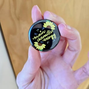 May include: A black button with a yellow sunflower design and the text "main character energy" in yellow script.