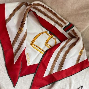 May include: A square silk scarf featuring a white center with a large gold horse bit graphic and the Gucci logo. The design includes brown leather belt motifs and a wide red border with green and red striped accents along the edges of the fabric.