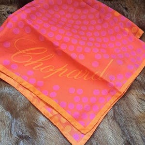 May include: A square silk scarf in a vibrant orange hue featuring a repeating pattern of small, bright pink polka dots. The word Chopard is printed in a large, elegant, cursive script across the center of the fabric in a matching orange tone.