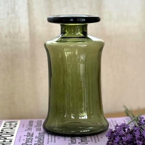 Può includere: Un vaso di vetro verde oliva con bordo nero, appoggiato su una pila di libri colorati. I libri sono etichettati "REPORTAGEN" e sono impilati. Un piccolo mazzo di fiori viola poggia sui libri.