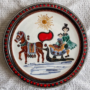 May include: A round ceramic plate with a winter scene. The plate features a horse-drawn sleigh with two figures, a sun, and a decorative border with red accents. The plate is brown, white, red, blue, and green.