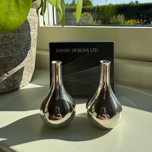 Puede incluir: Dos jarrones plateados en forma de lágrima con cuellos estrechos. Los jarrones son reflectantes, reflejando el entorno. Una caja negra con el texto "DANSK DESIGNS LTD" está detrás de ellos. Una planta verde es visible a la izquierda.