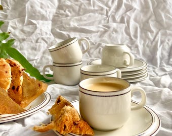 May include: A set of white ceramic coffee cups and saucers with a dark brown trim. The cups are stacked and arranged with plates and pastries on a white cloth background. One cup is filled with coffee.