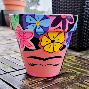 Puede incluir: Una maceta de terracota pintada con un diseño floral colorido y un retrato estilizado de Frida Kahlo. La maceta es rosa con contornos negros y presenta una flor amarilla, una flor azul y una flor rosa.