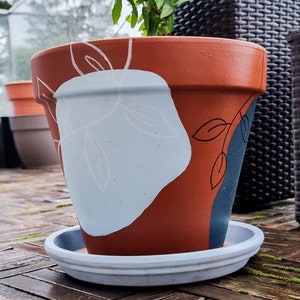 Könnte beinhalten: Ein Terrakotta-Blumentopf mit einem abstrakten Design in Weiß und Blau. Der Topf hat einen weißen Untersetzer und steht auf einem Holzdeck.