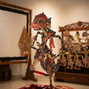 Könnte beinhalten: Eine javanische Schattenfigur, oder Wayang Kulit, mit komplexen Ausschnitten, bemalt in Schwarz, Rot und Gold. Die Figur ist auf einem zentralen Holzstab montiert und steht auf einem rechteckigen Holzsockel, der mit dunkelrotem Stoff bedeckt ist.
