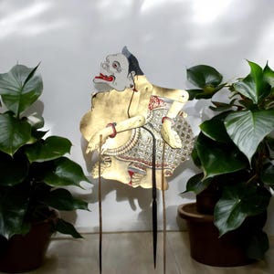 Könnte beinhalten: Eine traditionelle indonesische Schattenpuppe, oder Wayang Kulit, die eine mythische Kreatur mit einem grimmigen Ausdruck darstellt. Die Puppe ist aus Leder gefertigt und in Gold, Rot, Weiß und Schwarz bemalt. Sie ist auf Holzstöcken montiert, um in Schattenpuppenspielen verwendet zu werden.