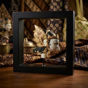 Puede incluir: Una escultura de cabeza de marioneta de madera con rostro blanco y detalles pintados en negro y oro, que incluye un tocado ornamentado y pendientes dorados colgantes, montada sobre una varilla central en un marco negro cuadrado.