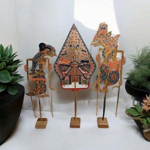 Puede incluir: Tres títeres de sombras tradicionales indonesios, o wayang kulit, en soportes de madera. Los títeres están hechos de cuero y pintados en colores vibrantes, incluyendo rojo, naranja, amarillo, azul y negro. Los títeres representan criaturas y figuras míticas.