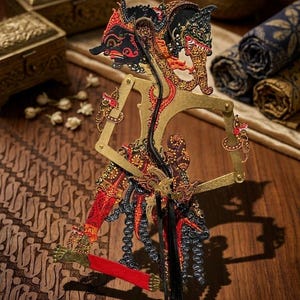 Op de afbeelding: Een traditionele Javaanse schaduwpop, bekend als wayang kulit, met ingewikkelde uitgesneden details. De figuur is beschilderd in zwart, rood en goud, met beweegbare armen bevestigd aan dunne staven voor beweging, voorzien van sierlijke patronen.