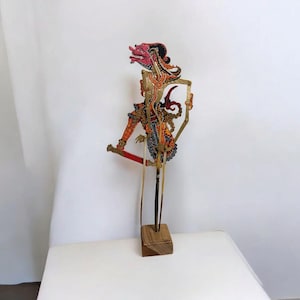 Puede incluir: Un títere de sombras tradicional indonesio, o wayang kulit, que representa una criatura mítica con un diseño rojo, naranja y negro. El títere está hecho de cuero y está montado en un soporte de madera.
