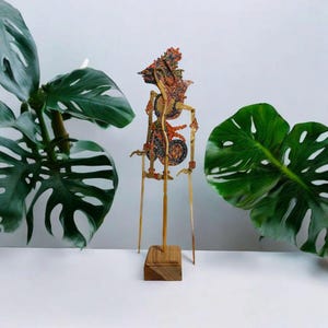 Puede incluir: Un títere de sombras tradicional indonesio, o wayang kulit, que representa una criatura mítica. El títere está hecho de cuero tallado y pintado de forma intrincada y está montado en un soporte de madera con varillas de bambú.
