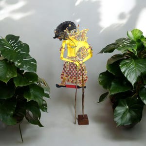 Puede incluir: Un títere de sombras tradicional indonesio, o wayang kulit, que representa a un personaje con un traje amarillo y dorado, cabello negro y una falda con estampado rojo y negro. El títere está montado en un soporte de madera con dos palos largos.