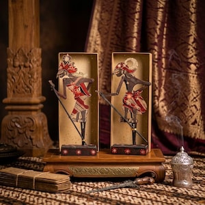 May include: Two Javanese wayang kulit shadow puppets displayed in individual vertical boxes on a wooden stand. The puppets are crafted with intricate perforated details, featuring red and gold accents, and are mounted on thin black rods.