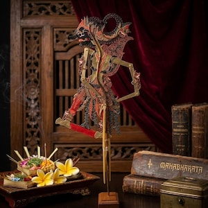 May include: An Indonesian Wayang Kulit puppet with detailed cut-out patterns in red, black, and gold. The puppet is displayed on a wooden stand. Incense and flowers are in the foreground, with antique books and a decorative box in the background.