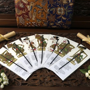 Può includere: Una collezione di segnalibri indonesiani Wayang Kulit, ciascuno con un design unico del personaggio. I segnalibri sono disposti a ventaglio su una superficie di legno, con una pochette fantasia e una pergamena nelle vicinanze. I burattini sono in tonalità di verde, oro e marrone.