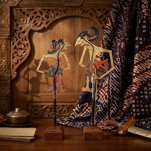 Op de afbeelding: Twee Indonesische Wayang Kulit poppen op houten sokkels. De poppen hebben gouden, rode en zwarte details. Ze staan tegen een achtergrond van gesneden hout en bedrukte stof, wat een traditionele uitstraling creëert. De poppen zijn ongeveer 30 cm hoog.