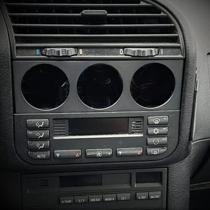 Peut inclure: Tableau de bord de voiture noir avec des bouches d'aération, trois ouvertures circulaires pour jauges et des boutons de commande de climatisation. Le tableau de bord comprend des boutons pour la température, la vitesse du ventilateur et d'autres fonctions du véhicule.