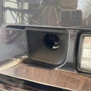 May include: Close-up of a black plastic bumper with a rectangular cutout. The cutout has a round hole in the centre.