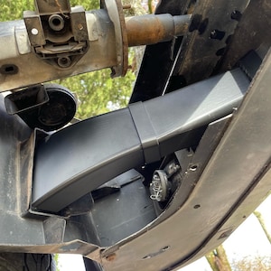 Op de afbeelding: Een schuin, zwart auto-onderdeel is op een voertuig gemonteerd. De afbeelding toont een close-up van de voorkant van het voertuig, met het onderdeel bevestigd aan de bumper en andere mechanische onderdelen. De achtergrond bevat een wazig zicht op bomen.