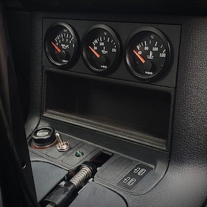 Puede incluir: Primer plano de un salpicadero de coche con tres indicadores negros que muestran la presión del aceite, el nivel de combustible y la temperatura. Debajo de los indicadores hay un encendedor de cigarrillos, un interruptor basculante y otros controles. El salpicadero es negro.