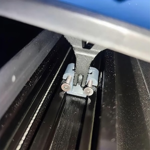 Puede incluir: Primer plano de un componente mecánico con un soporte de metal gris y dos piezas de plástico gris. Se ven dos tornillos plateados, junto con un marco negro y un acento azul.