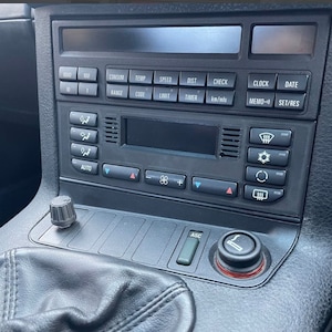 Può includere: Un cruscotto di un'auto nero con un display digitale, pulsanti e controlli. Il display mostra il testo "1000", "100", "CONSUM", "TEMP", "SPEED", "DIST", "CHECK", "CLOCK", "DATE", "10", "RANGE", "CODE", "LIMIT", "TIMER", "km/mis", "MEMO)" e "SET/RES".