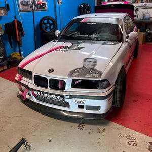 May include: A white BMW 3 series with pink and black racing stripes, a German Style sticker, and a GT21 sticker. The car has a sticker of a man with the text "Art & Creation" and "Plaque & Signal&eacute;tique Rallye".
