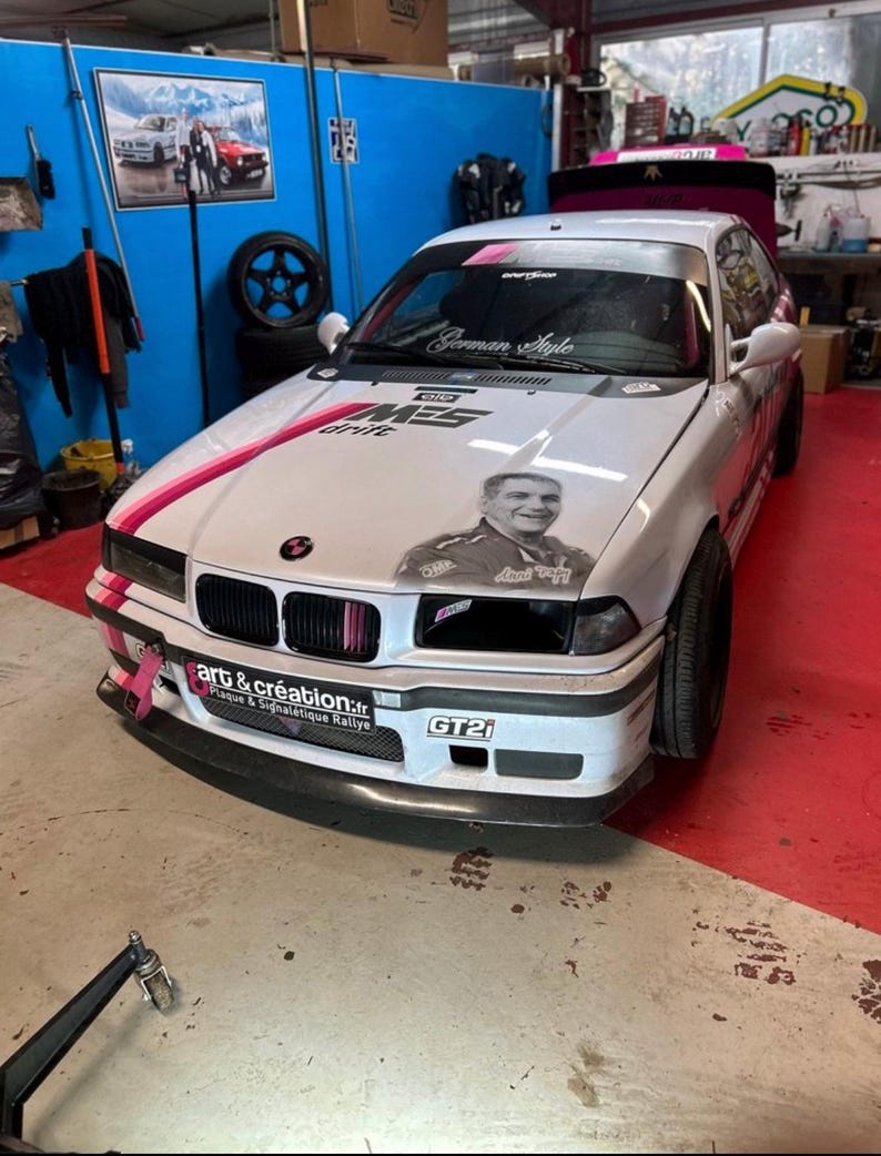 May include: A white BMW 3 series with pink and black racing stripes, a German Style sticker, and a GT21 sticker. The car has a sticker of a man with the text "Art & Creation" and "Plaque & Signal&eacute;tique Rallye".