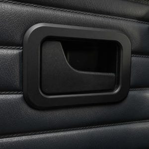 Puede incluir: Una manija de puerta interior negra insertada en un panel de cuero azul oscuro con costuras horizontales. La manija presenta un marco rectangular que rodea un mecanismo de palanca empotrado, integrado en la tapicería de la puerta.