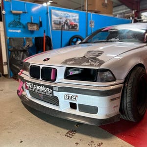 May include: A white BMW E36 with a black front bumper and a black and white graphic on the hood. The car has a pink tow strap and a "GT2i" sticker on the front bumper. The car is in a garage with a blue wall in the background.