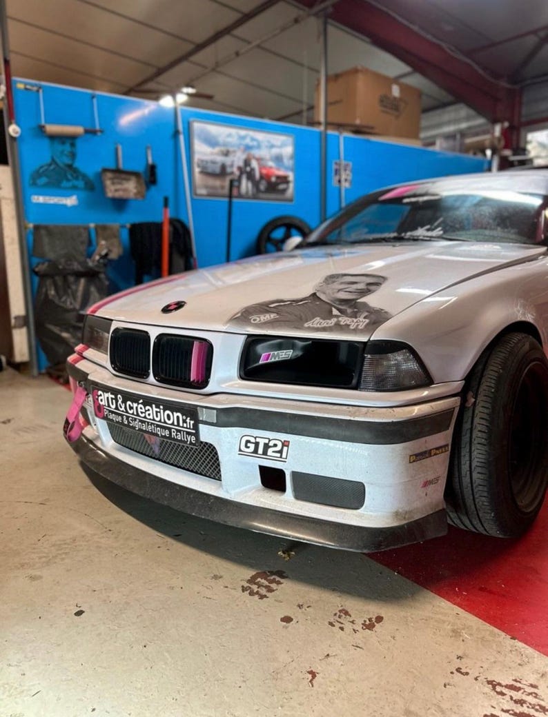 May include: A white BMW E36 with a black front bumper and a black and white graphic on the hood. The car has a pink tow strap and a "GT2i" sticker on the front bumper. The car is in a garage with a blue wall in the background.