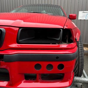 May include: A red car with black headlights and a black bumper. The car has three round holes in the bumper.