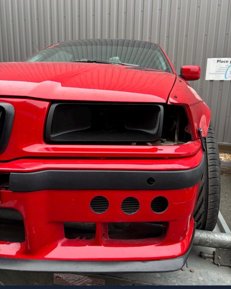 May include: A red car with black headlights and a black bumper. The car has three round holes in the bumper.