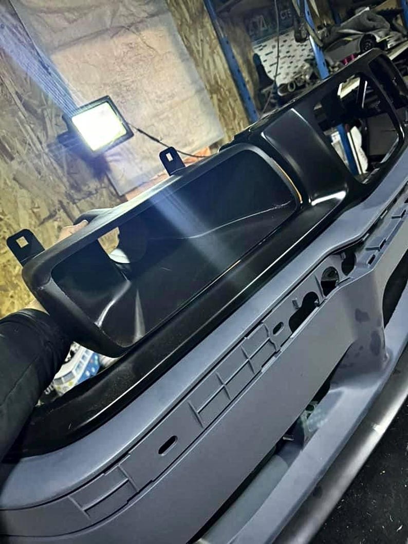 May include: A black plastic car bumper with a large black plastic grille. The bumper is painted a light gray color.
