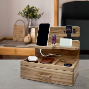 Op de afbeelding: Een houten organizer met een lade, twee planken en vakken om apparaten op te laden. Het heeft een oplaadkabel, een telefoon, een horloge, een kristal en een draadloze oplader. De organizer is gemaakt van licht hout met een natuurlijke afwerking.