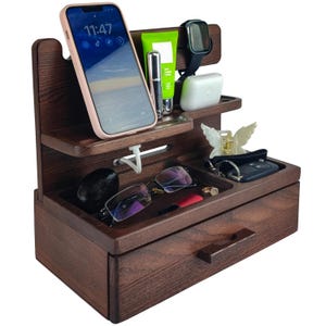 Könnte beinhalten: Ein dunkelbrauner Holzorganizer mit einer Schublade, mehreren Fächern und einem Telefonständer. Der Organizer verfügt über ein Ladekabel für das Telefon, eine Uhr, eine Brille, einen Schlüsselanhänger und eine kleine Flasche Lotion.