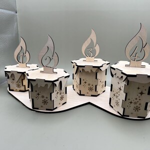 May include: Four wooden candle holders with numbers 1 through 4 on them. The holders are arranged in a row on a wooden base. Each holder has a snowflake design.