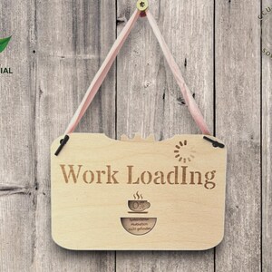 以下が含まれることがあります： 「Work Loading」の文字が刻まれた明るい茶色の木製サイン。コーヒーカップのグラフィックとローディングシンボルがあります。ピンクのリボンで吊り下げられ、木製の背景に配置されています。