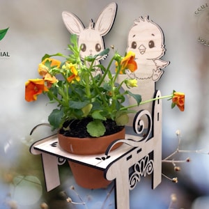 Puede incluir: Jardinera de madera con forma de banco, con diseño de conejito y pollito. Contiene una maceta de terracota con flores naranjas y amarillas. Incluye licencia comercial.