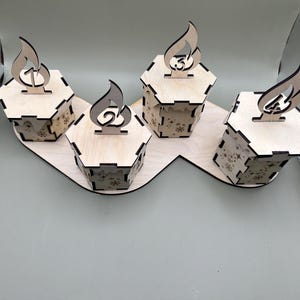May include: Four wooden advent calendar boxes with a flame design and numbers 1 through 4. The boxes are arranged in a curved line on a wooden base.