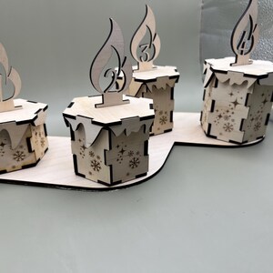 May include: Four wooden candle holders with a snowflake design. Each holder has a number on it, 1, 2, 3, and 4. The holders are arranged in a semi-circle on a wooden base.